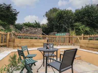 An outdoor patio area with a round table, chairs, wooden planter boxes, a picket fence, and greenery at Holly Cottage in West Coker