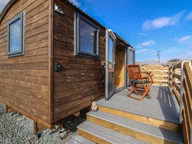 A wooden cabin with two windows and an open door leading to a deck with a wooden chair and table at Claerwen Nant-glas near Rhayader