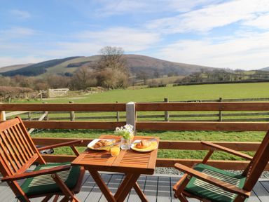 A wooden table with two croissants glasses of orange juice and a small flower vase on a patio with two wooden chairs overlooking fields and hills at Claerwen Nant-glas near Rhayader