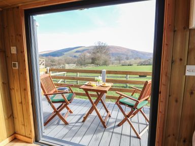 A balcony with two wooden chairs and a small table set with food and drinks overlooking fields and hills at Claerwen Nant-glas near Rhayader