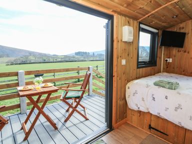A small wooden bedroom with a bed towels on it and an open door leading to a balcony with wooden chairs and a table with breakfast plates at Claerwen in Nant-glas near Rhayader