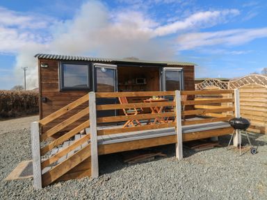 A small wooden cabin with open front doors a wooden deck with table and chairs and a grill on gravel at Claerwen in Nant glas near Rhayader