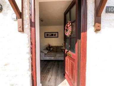 An open wooden door with a floral wreath leads to a room with a grey sofa and framed pictures on the wall at Chalk Cottage in Feltwell