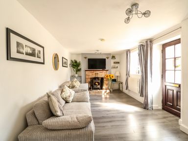 A living room with a sofa, pillows, a brick fireplace with a wood stove, a TV above the fireplace, a door, windows with curtains, a floor lamp, and framed pictures on the wall at Chalk Cottage in Feltwell