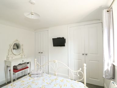 A bedroom with a white metal bed frame a floral bedspread a white vanity table with a mirror and a red cushioned stool a wall-mounted television and two white double door closets at Chalk Cottage in Feltwell