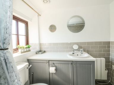 A bathroom with a sink countertop gray cabinets two round mirrors a window with plants and a toilet at Chalk Cottage in Feltwell