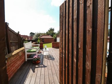A garden patio with wooden decking white bench with green cushions a small white table and potted plants at Chalk Cottage in Feltwell