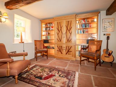 A room with two wooden chairs a beige armchair a wooden bookcase a guitar on a stand and a patterned rug on the floor at Eagle Farmhouse in Glenridding