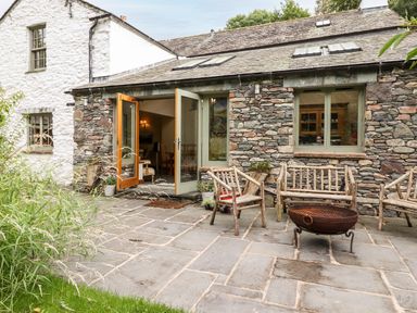 An outdoor stone patio with wooden chairs and a fire pit outside a stone cottage with green window frames at Eagle Farmhouse in Glenridding