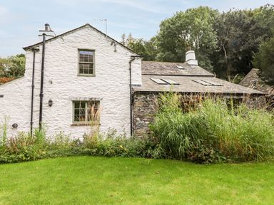 The exterior of a stone and white plaster house with windows a green lawn and tall plants at Eagle Farmhouse in Glenridding