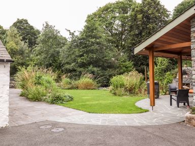 A garden area with green grass and bushes next to a covered patio with chairs at Eagle Farmhouse in Glenridding