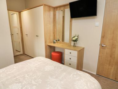 A bedroom with a bed a wooden wardrobe a dressing table with drawers a mirror a red stool and a wall mounted TV at The Warren in Hellifield