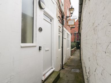 A narrow alleyway with white painted walls windows and a door on one side and a brick building with a hanging sign at 24 Baxtergate in Whitby