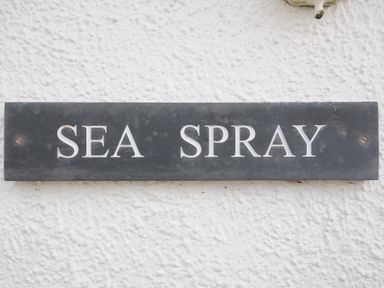 A black rectangular sign with white letters reading SEA SPRAY mounted on a white textured wall at Sea Spray in Port Isaac