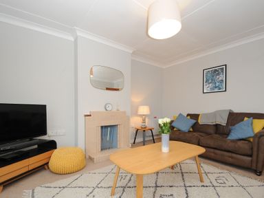 A living room with a brown sofa with blue and yellow cushions a light wooden coffee table with flowers a TV on a stand a fireplace and a lamp at Sea Spray Port Isaac