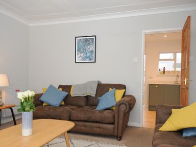 A living room with brown sofas blue and yellow cushions a wooden coffee table with white flowers and a doorway to a kitchen at Sea Spray in Port Isaac