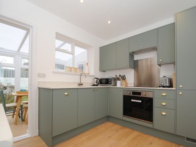 A kitchen with green cabinets, a built-in oven, microwave, kettle, toaster, and window looking into an adjoining room at Sea Spray in Port Isaac