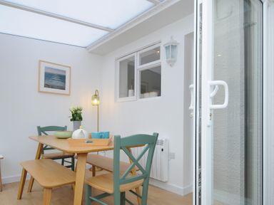 A dining area with a wooden table green chairs a bench a framed sea picture and a glass door at Sea Spray in Port Isaac