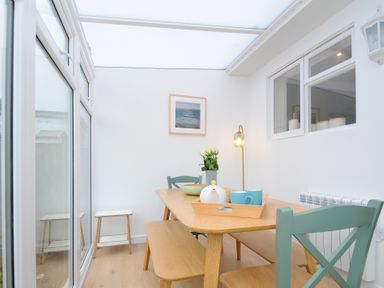 A dining area with a wooden table and bench a green chair a tray with cups and a teapot flowers and a framed picture on the wall at Sea Spray in Port Isaac
