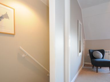 A hallway with stairs and a framed map on the wall next to a room with a dark chair and a round pillow at Sea Spray in Port Isaac