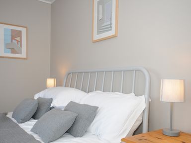 A bedroom with a bed with white and grey pillows bedside tables with lamps and framed pictures on the wall at Sea Spray in Port Isaac