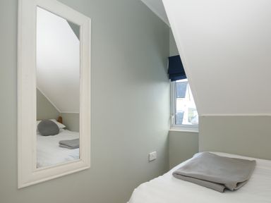 A small bedroom with a bed covered with a white sheet and a grey blanket on top a wall mirror reflecting another bed with pillows and a window with a dark blind