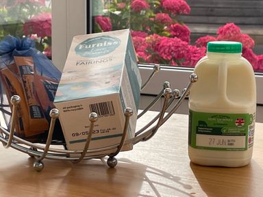 A kitchen table with a metal fruit bowl containing a box of Furniss Original Cornish Fairings and coffee sachets next to a bottle of semi skimmed milk with pink flowers visible outside the window at Sea Spray in Port Isaac