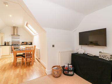 A dining area with a wooden table and chairs next to a kitchen with appliances and a skylight window and a living area with a flat screen tv mounted above a covered cabinet and baskets on the floor at Bayview Loft in Weymouth