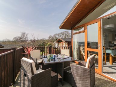 An outdoor deck with a table and four chairs next to a wooden building with glass doors at Meadow Lodge in Newton Ferrers