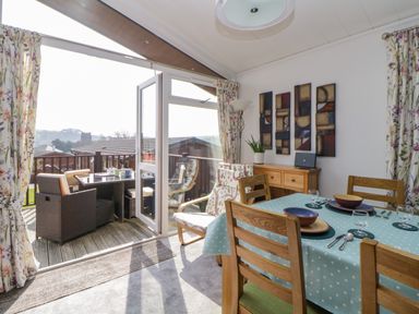 A dining area with wooden chairs and a table set with plates and glasses next to a door opening to an outdoor seating area at Meadow Lodge in Newton Ferrers