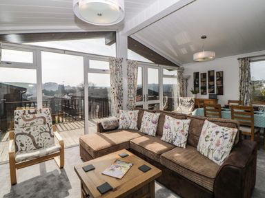 A living room with a sectional sofa and armchair near glass doors and a dining table with chairs at Meadow Lodge in Newton Ferrers