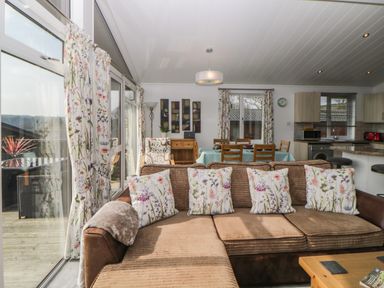 A living room with a brown sectional sofa and floral cushions next to large windows with floral curtains at Meadow Lodge in Newton Ferrers