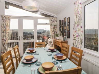 A dining room with a table set for six and wooden chairs next to glass doors and floral curtains at Meadow Lodge in Newton Ferrers