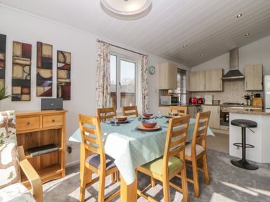 A dining area with a table set for six and an open kitchen with appliances at Meadow Lodge in Newton Ferrers