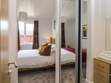A bedroom with a wooden bed and multicolored cushions near a window with patterned curtains at Meadow Lodge in Newton Ferrers