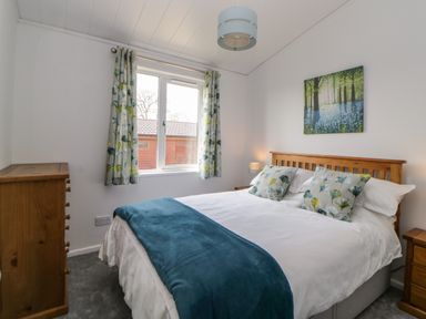 A bedroom with a double bed with floral pillows and blue blanket a wooden dresser and a window with floral curtains at Meadow Lodge in Newton Ferrers