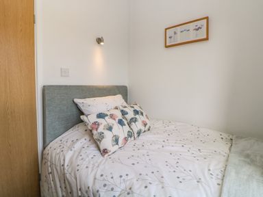 A bedroom with a bed with patterned bedding and two pillows under a wall light and a framed picture at Meadow Lodge in Newton Ferrers