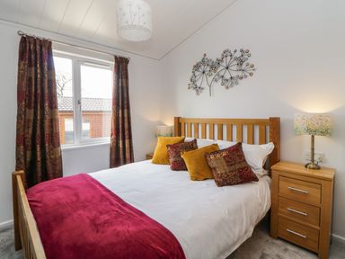 A bedroom with a wooden bed beside a window with curtain and a bedside table with lamp at Meadow Lodge in Newton Ferrers
