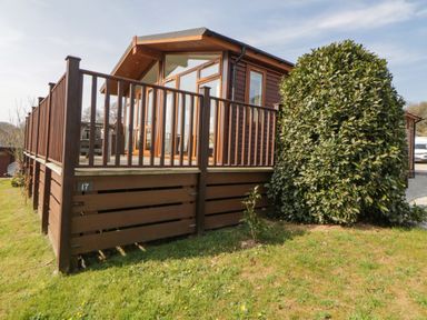 A wooden cabin with a raised deck and railing next to a large bush at Meadow Lodge in Newton Ferrers