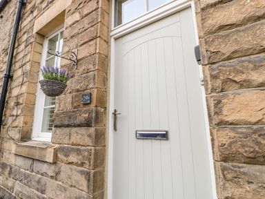 An exterior view of a stone house with a white door and window a hanging basket with purple flowers and a house number 29 at Toll House in Corbridge