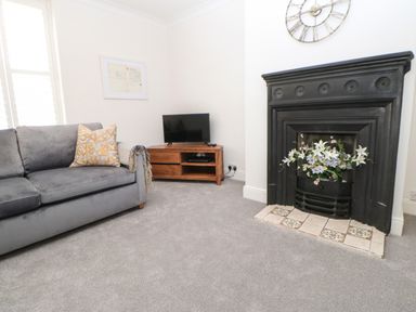 A living room with a gray sofa a wooden TV stand with a television and a black fireplace with flowers at Toll House in Corbridge