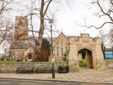A stone church with a clock tower behind a low stone wall and an arched stone entrance with paved walkway and benches in front at Toll House in Corbridge