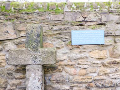 An old stone cross mounted on a stone wall with a blue plaque describing its history at The Old Market Cross in Corbridge