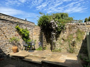An outdoor stone patio with two chairs a table climbing plants on stone walls and a wooden trellis at Toll House in Corbridge