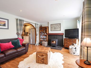 A living room with a leather sofa and colorful pillows a wooden chest on a rug a stove fireplace a bookshelf a tv on a dresser and curtains at The Hideout in Portree