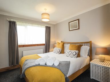 A bedroom with a wooden bed with white sheets yellow and gray pillows three rolled white towels on the bed a window with curtains and a bedside table with a lamp at The Hideout in Portree
