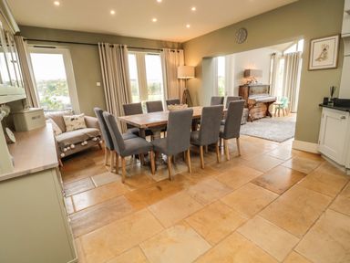 A dining room with a wooden table and eight grey chairs next to windows and a piano at Fontburn Farmhouse in Ewesley near Rothbury