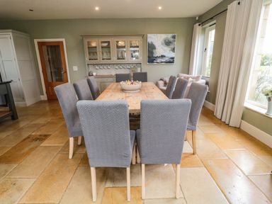 A dining room with a wooden table and eight grey chairs a sideboard with dishes and cups a painting and large windows with curtains at Fontburn Farmhouse in Ewesley near Rothbury