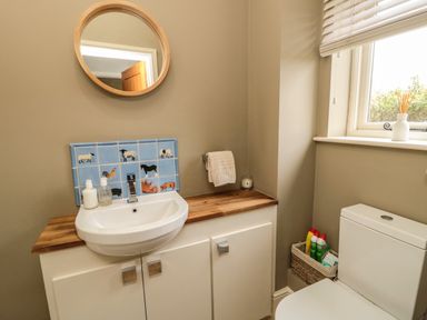 A bathroom sink with animal tiles below a round mirror a white toilet and a window with a vase on the sill at Fontburn Farmhouse in Ewesley near Rothbury