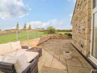 An outdoor patio with stone flooring a wicker sofa with cushions planter pots and a stone wall of a house at Fontburn Farmhouse in Ewesley near Rothbury
